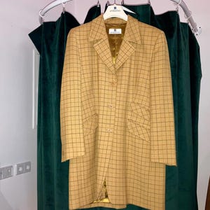 May include: A tan and black checked coat with a notched collar and two front pockets. The coat is suspended on a white hanger bearing the Givenchy logo. The coat is buttoned and has a gold lining. It hangs before a green curtain.