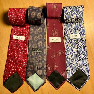 Könnte beinhalten: Vier Krawatten mit verschiedenen Mustern und Farben werden gezeigt. Eine ist rot mit weißen Punkten, eine weitere ist dunkelgrau mit einem floralen Design, und zwei sind bordeauxfarben mit geometrischen Mustern. Die Krawatten tragen das Label "BOSS".