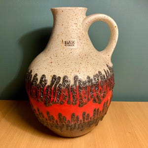 May include: A ceramic pitcher with a handle, featuring a speckled beige upper half and a vibrant red lower half with a textured, lava-like design. The pitcher has a label that reads "EIAX KIRAVA".