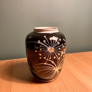 以下が含まれることがあります： 白と金のフラワーデザインが施された黒いセラミックの花瓶。花瓶は丸いボディと細い首をしています。