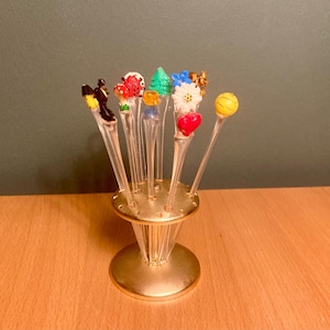 Op de afbeelding: Een set decoratieve cocktailprikkers met kleurrijke toppers, waaronder een lieveheersbeestje en een bloem, gepresenteerd in een goudkleurige houder. De prikkers zijn transparant met diverse ontwerpen. De houder heeft een ronde basis en een uitlopend, geribbeld lichaam.