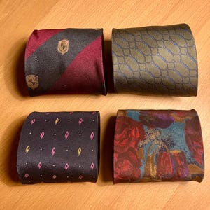 May include: Four rolled neckties. One is burgundy and black striped with a gold crest. Another is olive green with a blue chain pattern. The third is black with pink diamond shapes. The last has a floral pattern.