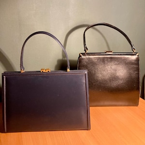Puede incluir: Dos bolsos vintage. Uno es azul marino con cierre y enganches dorados. El otro es negro con cierre y enganches dorados. Ambos tienen forma rectangular y asas superiores.