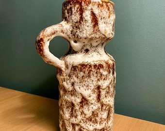 May include: A brown and white ceramic vase with a textured surface and a single handle. The vase has a narrow neck and a wider base.