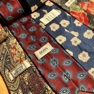 May include: A collection of various patterned neckties. The ties feature designs such as floral, paisley, and geometric patterns in colors like navy, burgundy, and gold. Several ties have brand labels, including BOSS and GIANNI VERSACE.