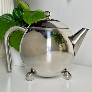 May include: A polished silver teapot with a spherical body, a curved handle, and a spout. The teapot rests on three small ball feet. The lid has a small knob. The teapot reflects the surrounding environment.
