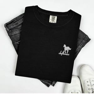 May include: Black crew neck t-shirt with a white horse graphic and the word "deftones" in a script font. The shirt is folded on top of a pair of dark gray denim jeans. A white tag is visible on the shirt. White sneakers are at the bottom.