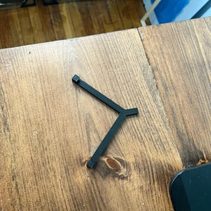 May include: A black, Y-shaped metal object with a square end on each of its three arms. The object is resting on a wooden surface with a visible wood grain.