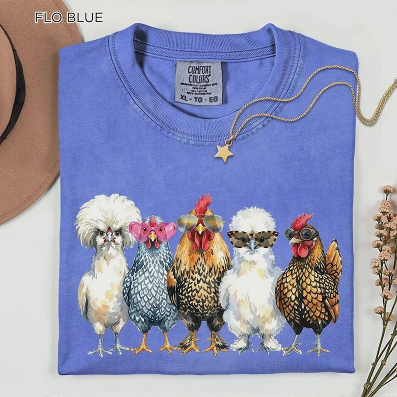 May include: A Flo Blue Comfort Colors t-shirt featuring a graphic of five chickens wearing various styles of sunglasses. The chickens are arranged in a row, each with distinct plumage patterns and colors, including white, grey, and brown feathers.