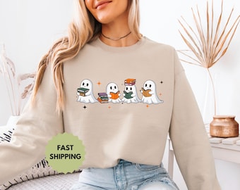 Puede incluir: Sudadera beige con una ilustración de fantasmas de dibujos animados leyendo libros. Los fantasmas son blancos con ojos negros y los libros son de varios colores. Estrellas naranjas y la inscripción "FAST SHIPPING".