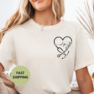 May include: Cream-colored t-shirt featuring a black heart-shaped design with a heartbeat line, stethoscope, and the name "Michelle." The shirt has short sleeves and a round neckline. A green and yellow sticker that says "FAST SHIPPING" is visible.