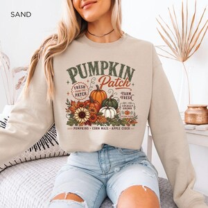 May include: A sand-colored crewneck sweatshirt featuring a graphic print with the text PUMPKIN PATCH, FRESH FROM THE PATCH, FARM FRESH, LOCALLY GROWN, and PUMPKINS CORN MAZE APPLE CIDER. The design includes illustrations of pumpkins, a sunflower, and leaves.