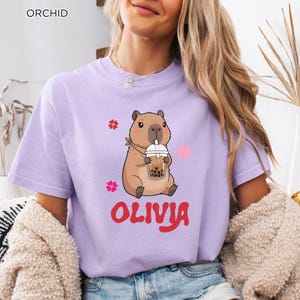 Puede incluir: Camiseta color orquídea con un carpincho de dibujos animados bebiendo té de burbujas. El carpincho lleva una bufanda roja y está rodeado de flores rosas y gráficos de corazón. El nombre "OLIVIA" está escrito en rojo debajo.