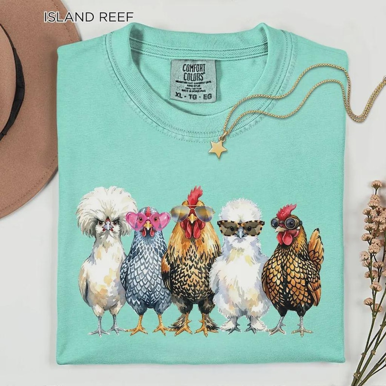 May include: A mint green crew neck t-shirt featuring a graphic of five chickens wearing various styles of sunglasses. The shirt has a Comfort Colors label at the neck with the text XL TG EG. A gold star pendant necklace rests on the fabric.