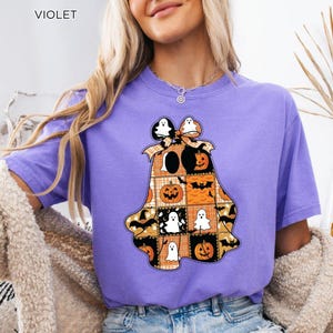 Może przedstawiać: Fioletowa koszulka z motywem Halloween. Wzór to patchworkowy duch z dyniami, nietoperzami i postaciami duchów. Słowo "VIOLET" jest wydrukowane w lewym górnym rogu.
