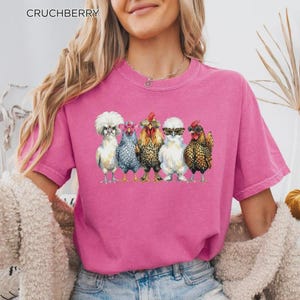 May include: A bright pink short-sleeved t-shirt featuring a graphic print of five chickens standing in a row. Each chicken wears different colored sunglasses. The text CRUCHBERRY is printed in the upper left corner of the image.