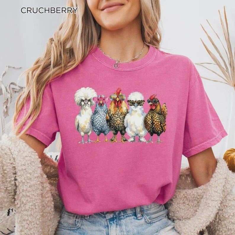 May include: A bright pink short-sleeved t-shirt featuring a graphic print of five chickens standing in a row. Each chicken wears different colored sunglasses. The text CRUCHBERRY is printed in the upper left corner of the image.
