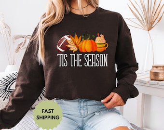 Puede incluir: Sudadera marrón oscuro con un diseño de otoño. El diseño incluye un balón de fútbol americano, hojas de otoño, una calabaza y una taza de café. El texto "'Tis the Season" está impreso en blanco.