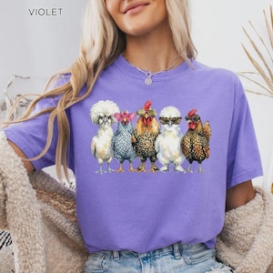 May include: A violet t-shirt featuring a graphic print of five chickens standing in a row. The chickens are wearing various styles of glasses, including heart-shaped, round, and aviator frames. The text VIOLET is printed in the upper left corner.