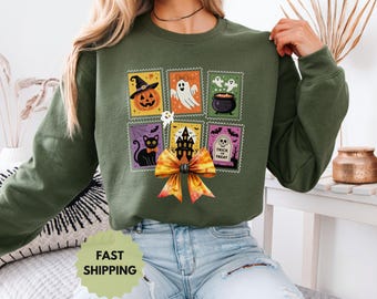 Può includere: Felpa verde scuro con un collage di disegni di francobolli a tema Halloween. I francobolli raffigurano una zucca, un fantasma, un calderone, un gatto nero, una casa infestata e una lapide con la scritta "Trick or Treat". Un grande fiocco arancione è sotto.