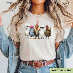 May include: An ivory t-shirt featuring a graphic print of five chickens standing in a row. Each chicken wears different colored glasses, including pink, blue, and black frames. The shirt has short sleeves and a crew neckline, presented in a neutral tone.