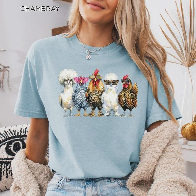 May include: A light blue t-shirt featuring a graphic print of five chickens wearing various styles of sunglasses. The text CHAMBRAY is printed in the upper left corner. The chickens are depicted in different colors including white, grey, and brown.