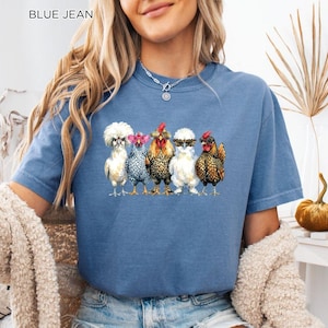 May include: A blue jean colored t-shirt featuring a graphic print of five chickens standing in a row. Each chicken wears different colored sunglasses, including pink, black, and leopard print frames. The text BLUE JEAN is printed in the top corner.