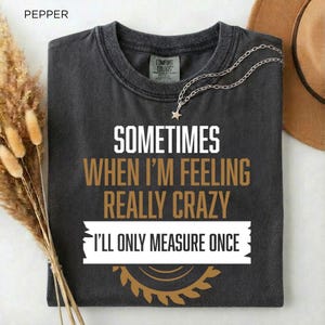 Op de afbeelding: Donkergrijs t-shirt met de tekst "SOMETIMES WHEN I'M FEELING REALLY CRAZY I'LL ONLY MEASURE ONCE". Het shirt heeft een ronde hals en een Comfort Colors-label. Een zilveren ketting met een sterhanger en een bruine hoed zijn ook zichtbaar.