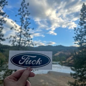 May include: A white sticker with a navy blue oval design, featuring the word "Fuck" in a stylized font, resembling the Ford logo. The sticker is held up against a backdrop of a river, mountains, and a cloudy sky.