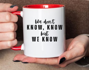 May include: White ceramic mug with a red interior and handle. The mug features the text "We don't KNOW, KNOW but WE KNOW" in black. The mug is held by a person wearing a striped sweater.