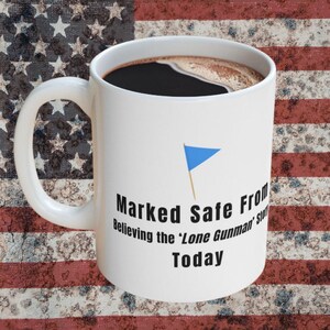 May include: A white ceramic coffee mug filled with dark coffee. The mug features the text "Marked Safe From Believing the 'Lone Gunman' Story Today" and a blue flag graphic. The background is an American flag.