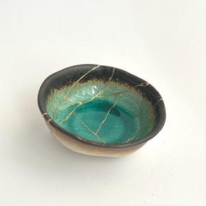 Könnte beinhalten: Kleine, handgefertigte Keramikschale mit einzigartiger Kintsugi-Reparatur. Die Schale hat einen türkisfarbenen Innenraum, einen grünen und schwarzen Farbverlauf am Rand und goldfarbene Linien, die die reparierten Risse hervorheben. Die Außenseite ist hellbraun.