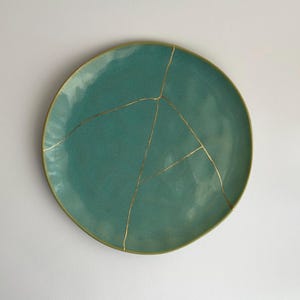 Puede incluir: Un plato de cerámica de color verde azulado con líneas de craquelado doradas. El plato es redondo y tiene un borde ligeramente irregular.