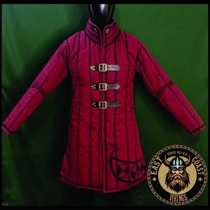 May include: A burgundy quilted coat with vertical stitching and three leather straps with buckles. The coat has long sleeves and a high collar. A circular logo with a Viking is on the lower right. The coat is designed for historical reenactment.
