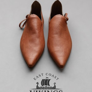 May include: A pair of tan leather slip-on shoes with pointed toes and side tie closures. Below the shoes is a black logo featuring a stylized Viking ship and the text EAST COAST VIKINGS, which includes a small flag and decorative knotwork.