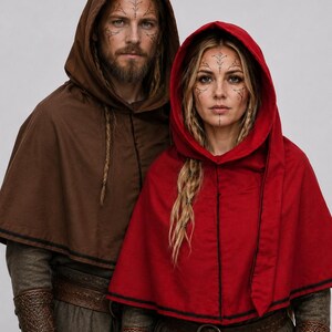 May include: Two people wearing hooded capes. One is brown, the other is red. Both have intricate tattoos on their faces and arms. They are also wearing long-sleeved shirts and belts. The capes have a black trim.