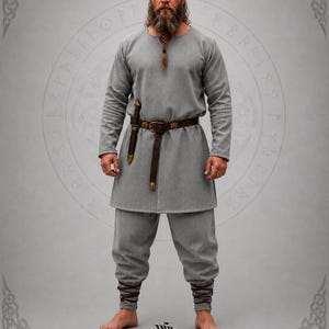 May include: A grey long-sleeved tunic and matching trousers set. The tunic features a round neckline and a brown leather belt with metal accents. The trousers are fitted at the ankles with brown leather wraps. The text EAST COAST VIKINGS is visible.