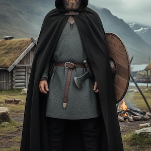May include: A person in a black hooded cloak, gray tunic, and black pants, holding a shield and axe. The person is standing in a grassy field with a small wooden building and mountains in the background. A campfire is visible.
