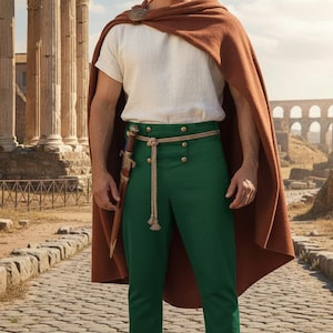 Op de afbeelding: Een persoon in een Romeins geïnspireerde outfit, met een bruine cape, een wit T-shirt en een groene broek. De outfit omvat een touwriem, een decoratieve broche en bruine sandalen. Een dolk is aan de riem bevestigd. De achtergrond toont oude ruïnes.