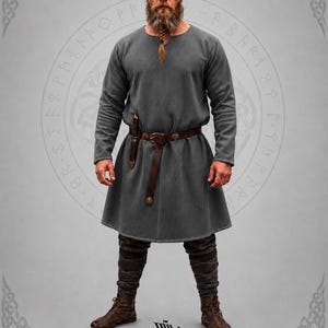 Puede incluir: Una túnica gris con mangas largas, un cinturón de cuero marrón y una funda para cuchillo. El atuendo incluye leggings y botas marrones. La imagen presenta el logotipo "East Coast Vikings".
