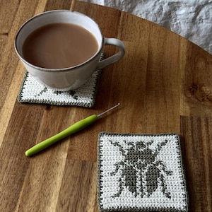May include: Two square crochet coasters featuring a white background with a dark olive green beetle motif centered on each. The coasters have a matching olive green border. A light green crochet hook lies diagonally between the two coasters.