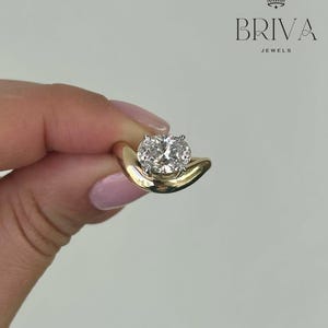 May include: A gold ring featuring a large, round, clear gemstone. The ring has a distinctive, curved design. The brand name "BRIVA JEWELS" is displayed in black text with a crown logo.
