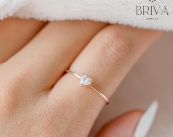 Op de afbeelding: Een delicate gouden ring met een hartvormige edelsteen. De ring is gezet op een slanke band en wordt gepresenteerd tegen een zachte, neutrale achtergrond. De sieraden zijn van het merk "BRIVA JEWELS".