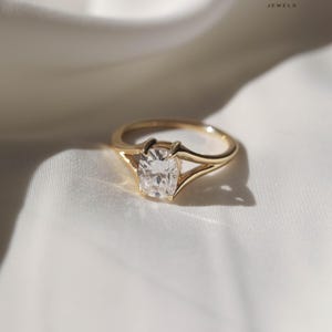 May include: A gold ring with a large, square-cut, clear gemstone. The ring has a delicate band and a unique setting that holds the gemstone securely. The ring is displayed on a white, textured surface. The logo "BRIVA JEWELS" is in the upper right corner.