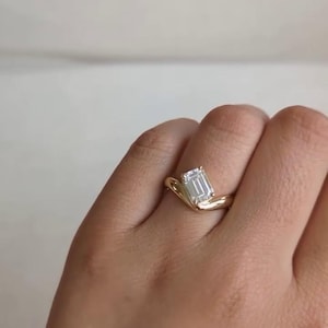 May include: A gold ring featuring an emerald-cut diamond. The ring has a distinctive, curved design.