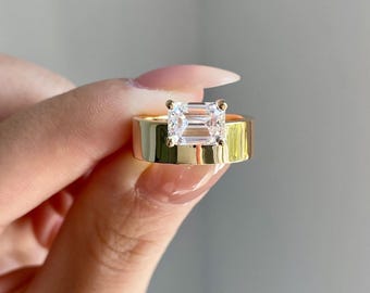 May include: A gold-colored ring with a large, rectangular, clear gemstone. The ring has a wide band and is held up by a hand. The brand name "BRIVA JEWELS" is visible in the upper right corner.