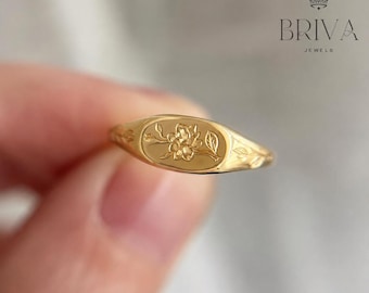 Può includere: Un anello con sigillo d'oro con un design floreale. L'anello presenta una faccia ovale con un motivo inciso di fiori e foglie. L'anello è di colore oro caldo ed è probabilmente in metallo.
