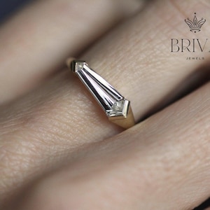 May include: A gold ring with a baguette-cut diamond. The diamond is set in a geometric design, and the ring is shown on a finger. The ring is from Briva Jewels.