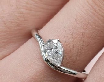 May include: A silver ring featuring a pear-cut diamond. The diamond is set in a flowing, curved band. The ring is displayed on a finger, highlighting the design and brilliance of the gem. The ring is from Briva Jewels.
