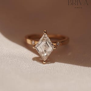 Könnte beinhalten: Ein goldener Ring mit einem großen, klaren, rautenförmigen Edelstein als Mittelpunkt. Der Ring hat kleine, eingebettete Steine und ist vor einem weichen, neutralen Hintergrund platziert. Der Markenname "BRIVA JEWELS" ist sichtbar.